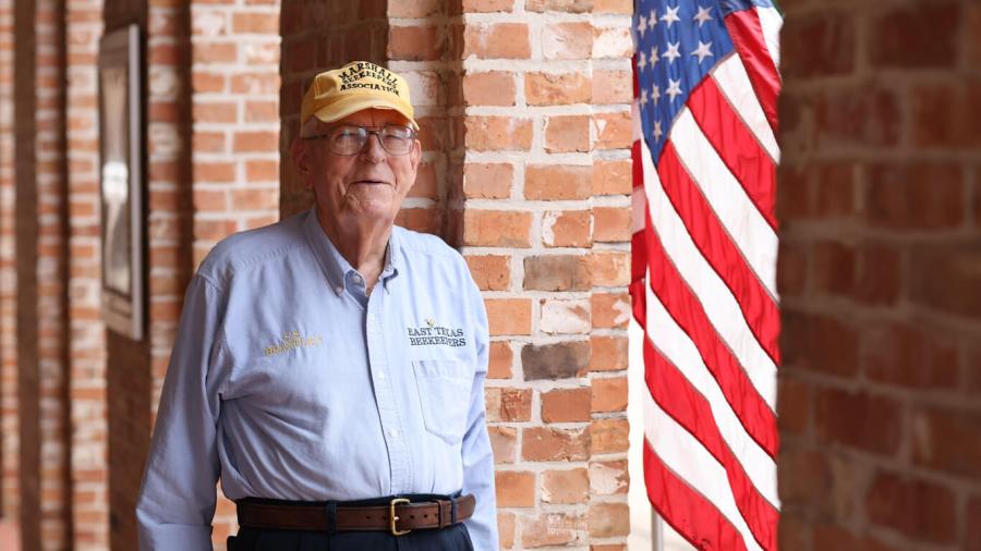 Stanford Brantley, clad in an East Texas Beekeepers shirt, stands against the red brick of Baylor University, with an American flag waving in the background